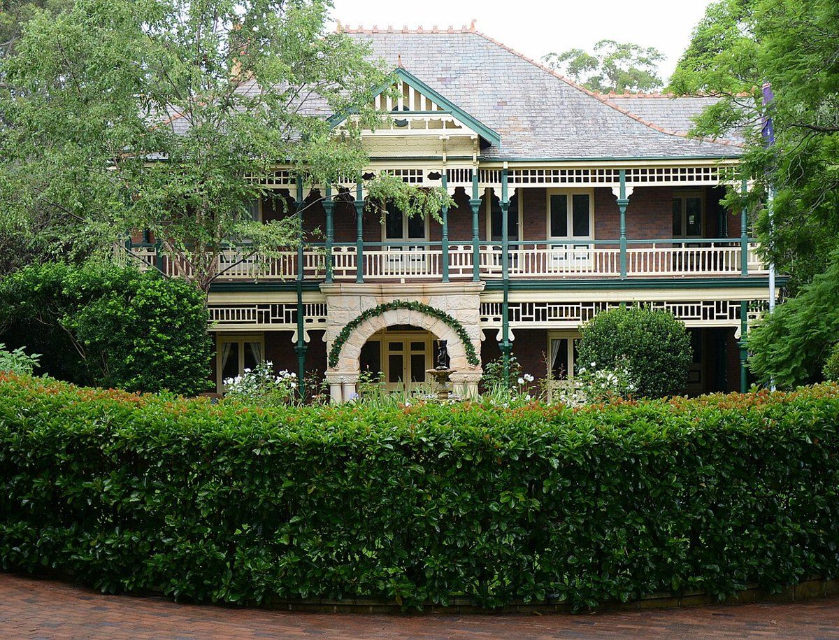 A large two-storey house with wide verandahs behind a tall clipped hedge in Ku-ring-gai Avenue, Turramurra